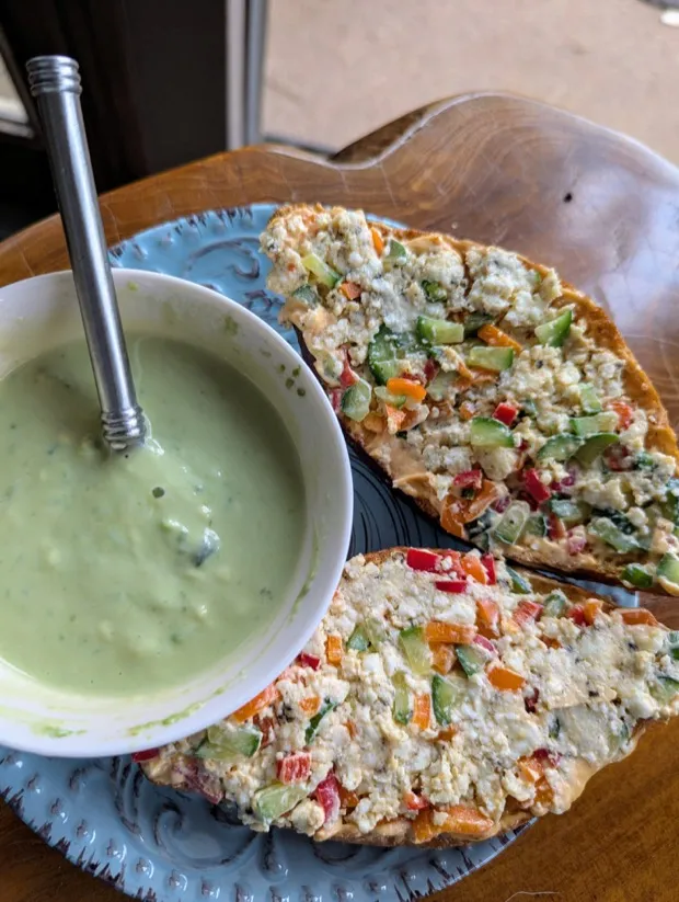 Two slices of sourdough spread with hummus and piled with a paneer scramble studded with diced cucumber and red and orange peppers, beside a bowl of green herb-yogurt dip